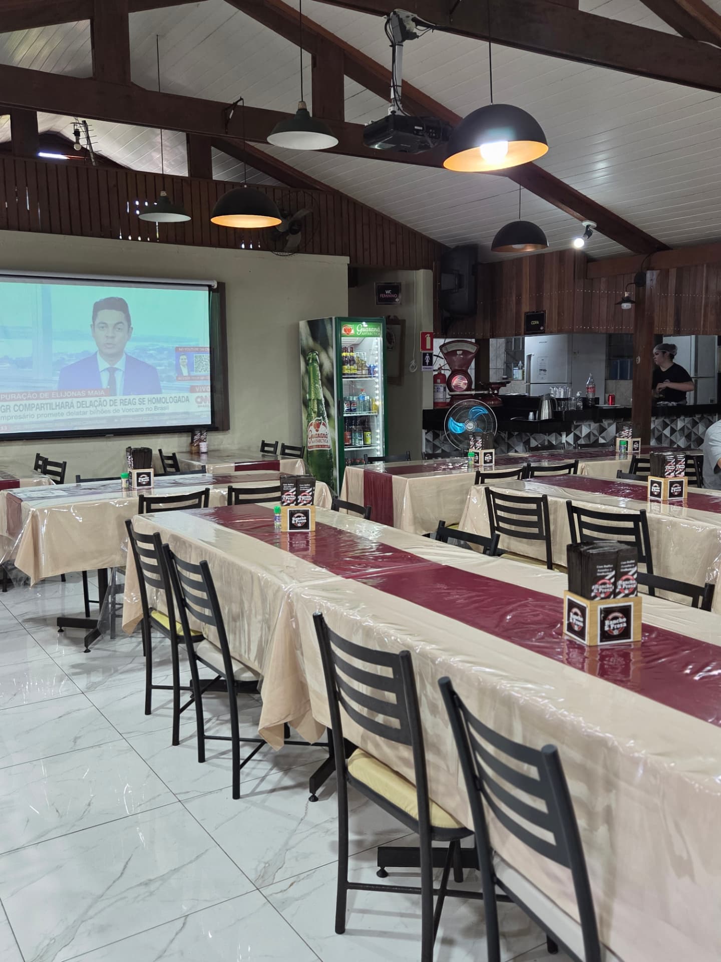 Salão interno do Rancho e Prosa com mesas compridas, telão e iluminação pendente